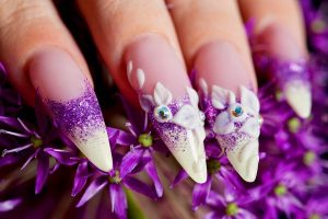 Long stiletto nails with purple glitter and 3D floral art.