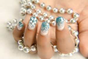 Short almond-shaped nails with a glittery blue and white gradient finish, floral accents, elegant and festive style, adorned with rhinestones.